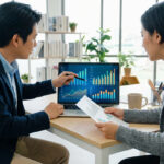 Deux professionnels analysent des graphiques sur un ordinateur portable. L'homme pointe l'écran, la femme tient des documents.