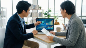 Deux professionnels analysent des graphiques sur un ordinateur portable. L'homme pointe l'écran, la femme tient des documents.